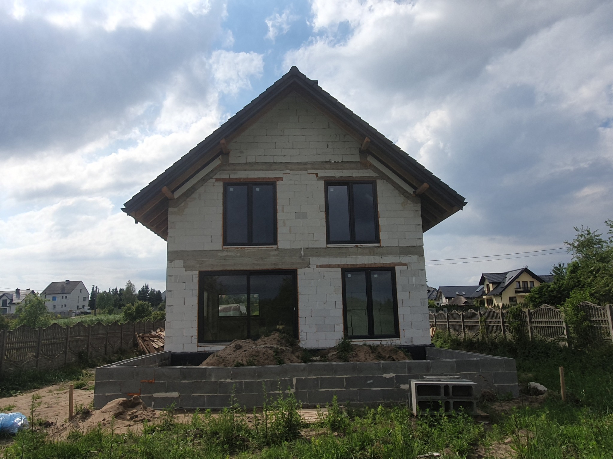 Dwukondygnacyjny dom w stanie surowym otwartym z czarną stolarką okienną, zbudowany z białych bloczków, na fundamencie z szarej kostki, w otoczeniu zieleni.