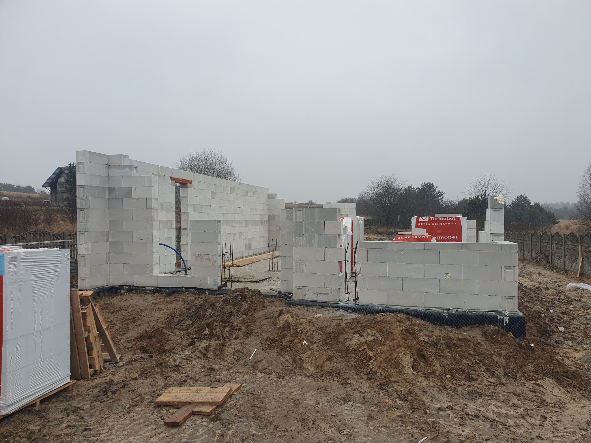 Wznoszenie ścian parterowego domu z betonu komórkowego Termobet na fundamencie, widoczne zbrojenia i paleta materiałów. Szary, pochmurny dzień, ziemiste podłoże.