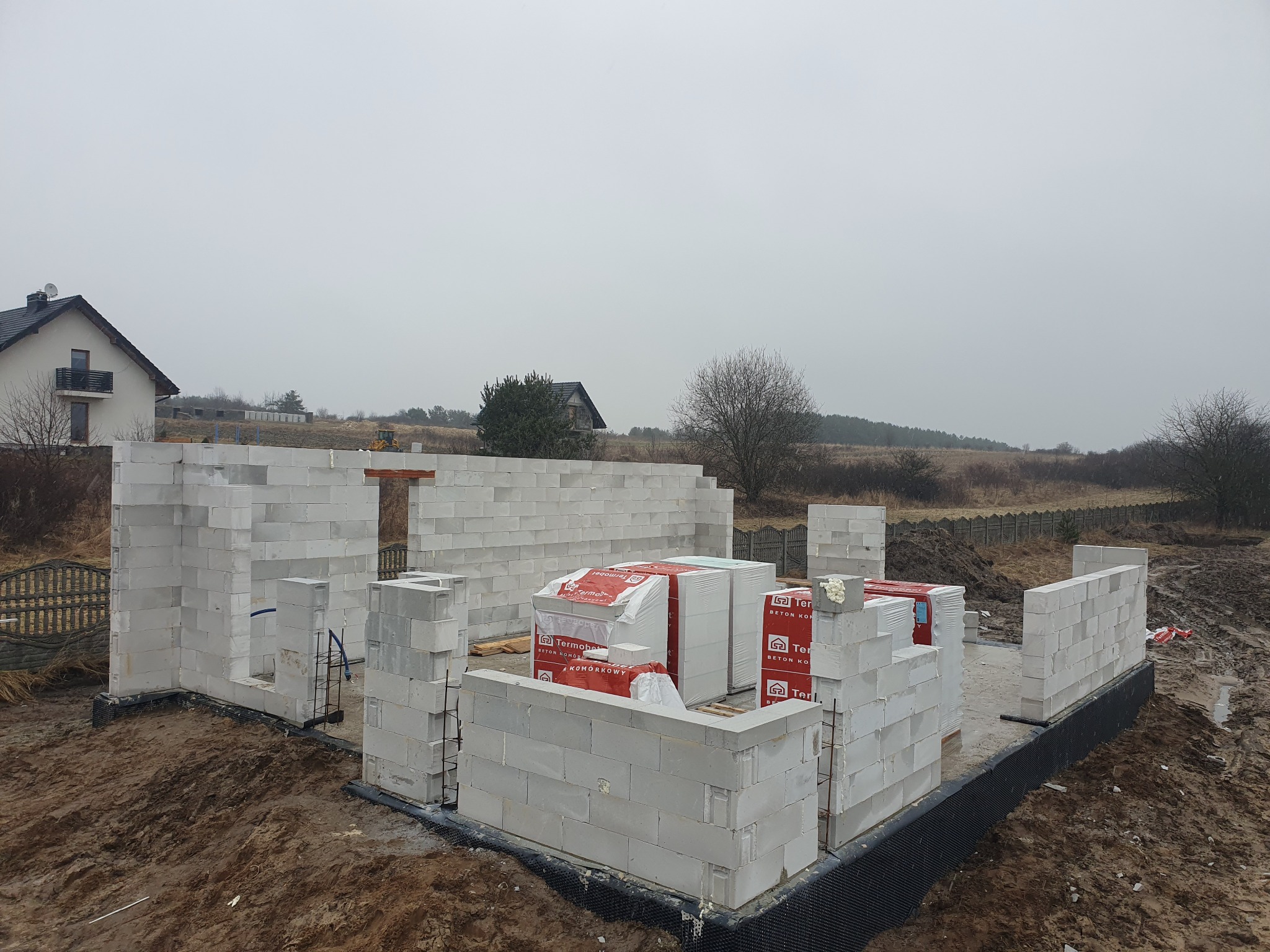 Budowa domu z betonu komórkowego: ściany parteru wzniesione, palety z bloczkami Termobet na fundamencie. Widok na posesję i pochmurne niebo.