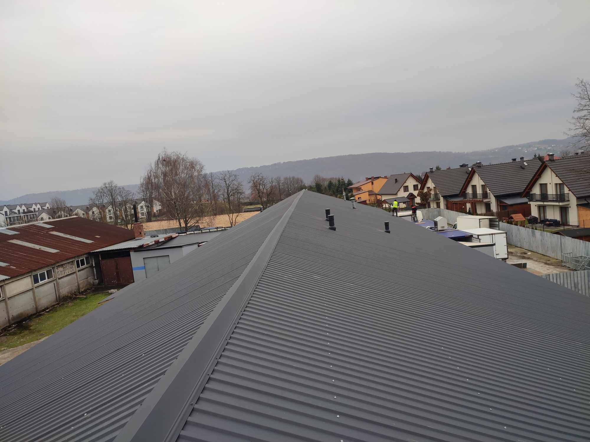 Szary, blaszany dach budynku z widocznymi kominami wentylacyjnymi na tle zabudowy miejskiej i zalesionych wzgórz w pochmurny dzień.