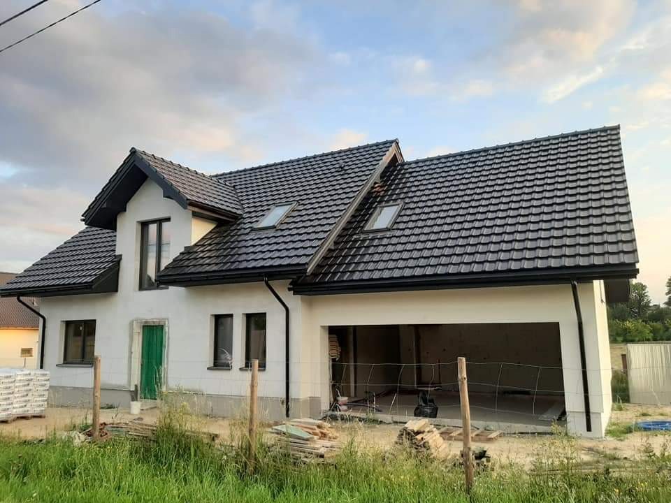 Nowoczesny dom jednorodzinny z ciemną dachówką i jasną elewacją, widoczny z zewnątrz, z otwartym garażem i zielonymi drzwiami wejściowymi. Częściowo wykończony.