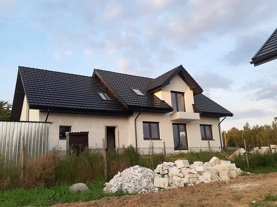 Niedokończony dom z ciemnym dachem i białymi ścianami. Widoczne sterty bloczków i niezabezpieczone otwory okienne. Nierówny teren i elementy ogrodzenia.