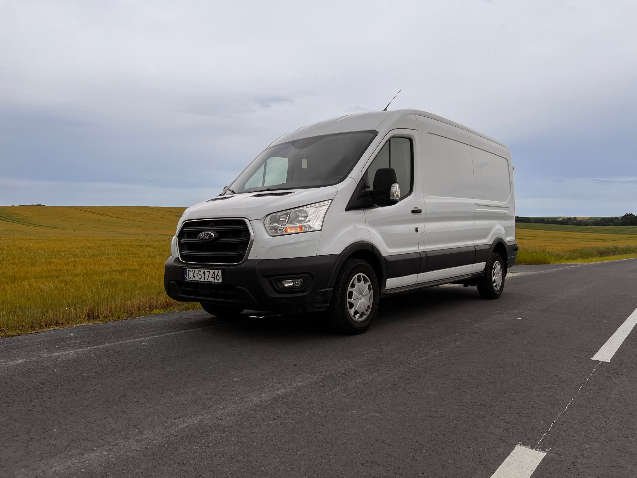 Biały Ford Transit na asfalcie z polem w tle. Rejestracja DX 51746. Zdjęcie z boku, pochmurne niebo.