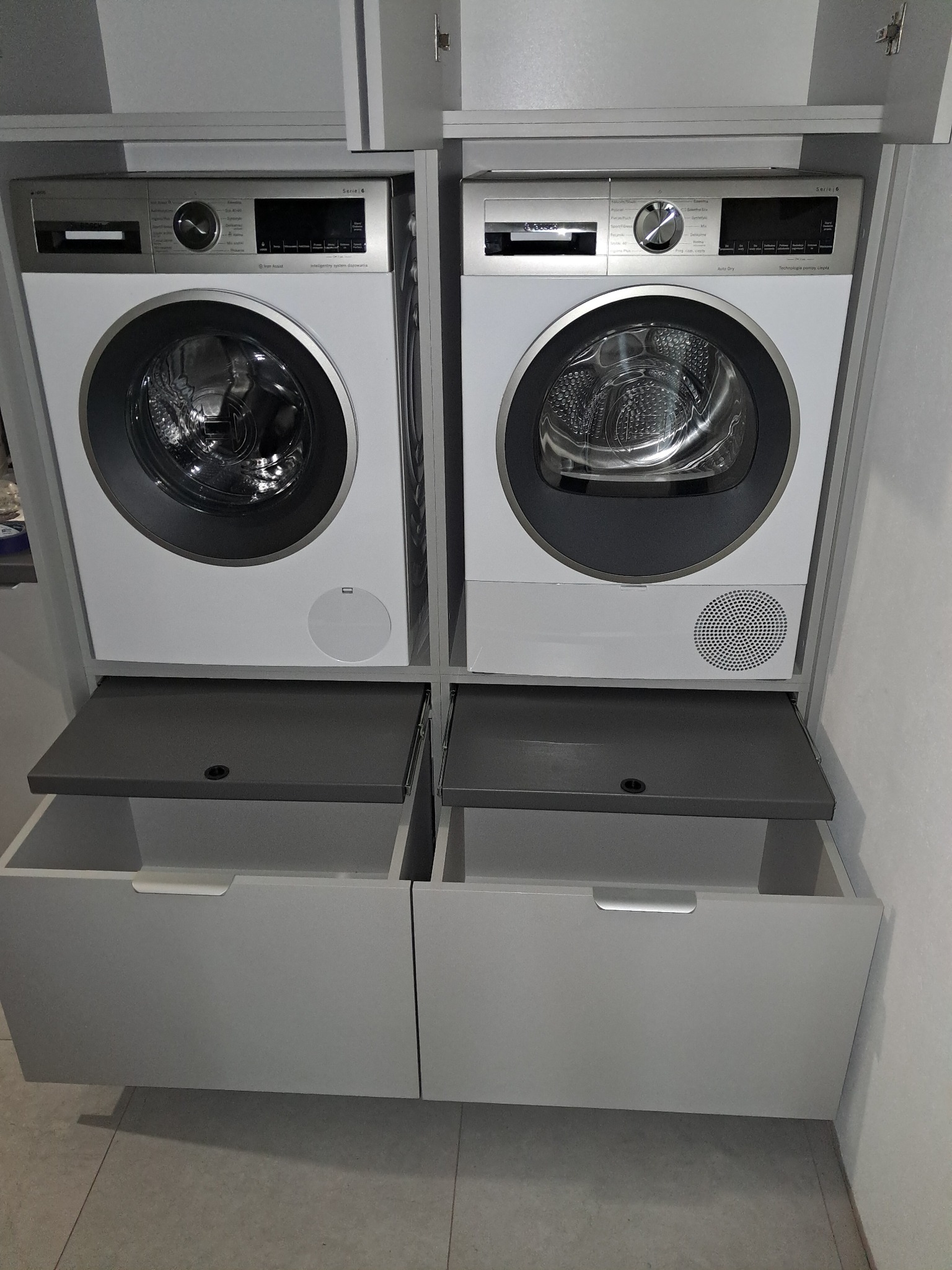 Szara zabudowa meblowa z pralką i suszarką marki Bosch Serie 6, z wysuwanymi półkami i szufladami. Nowoczesny design, minimalistyczne uchwyty.
