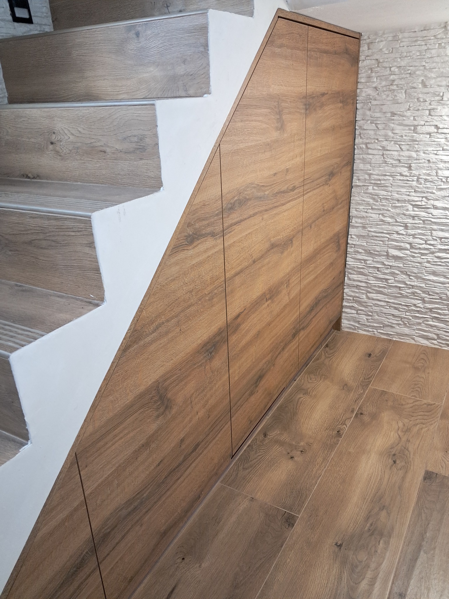 Drewniana szafa wnękowa pod schodami, dopasowana do koloru podłogi. Białe schody i dekoracyjna ściana z białego kamienia w tle.