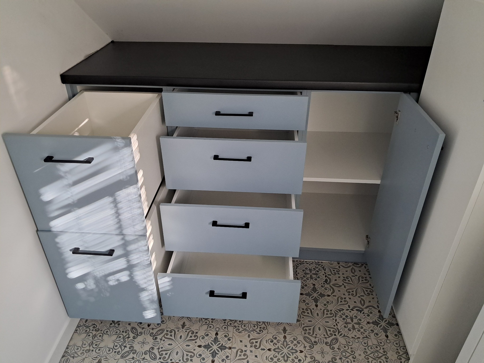 Niebieska szafa z otwartymi szufladami i drzwiczkami, czarne uchwyty, wzorzysta podłoga, zabudowa pod skosem, wnętrze szafy w kolorze białym.