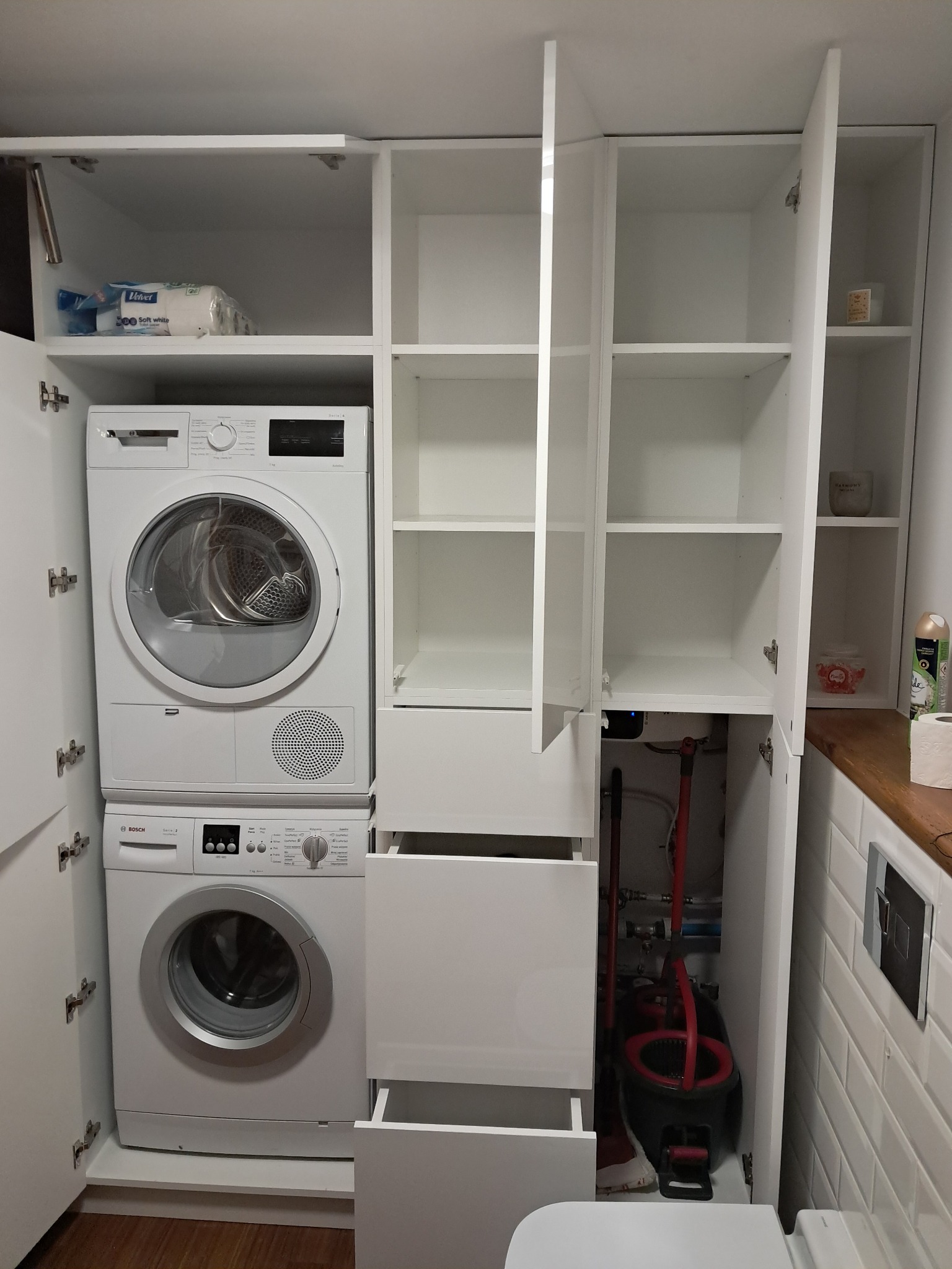 Biała szafa wnękowa w łazience z pralką i suszarką w zabudowie, otwarte półki i szuflady. Widoczne akcesoria łazienkowe i mop. Minimalistyczny design.