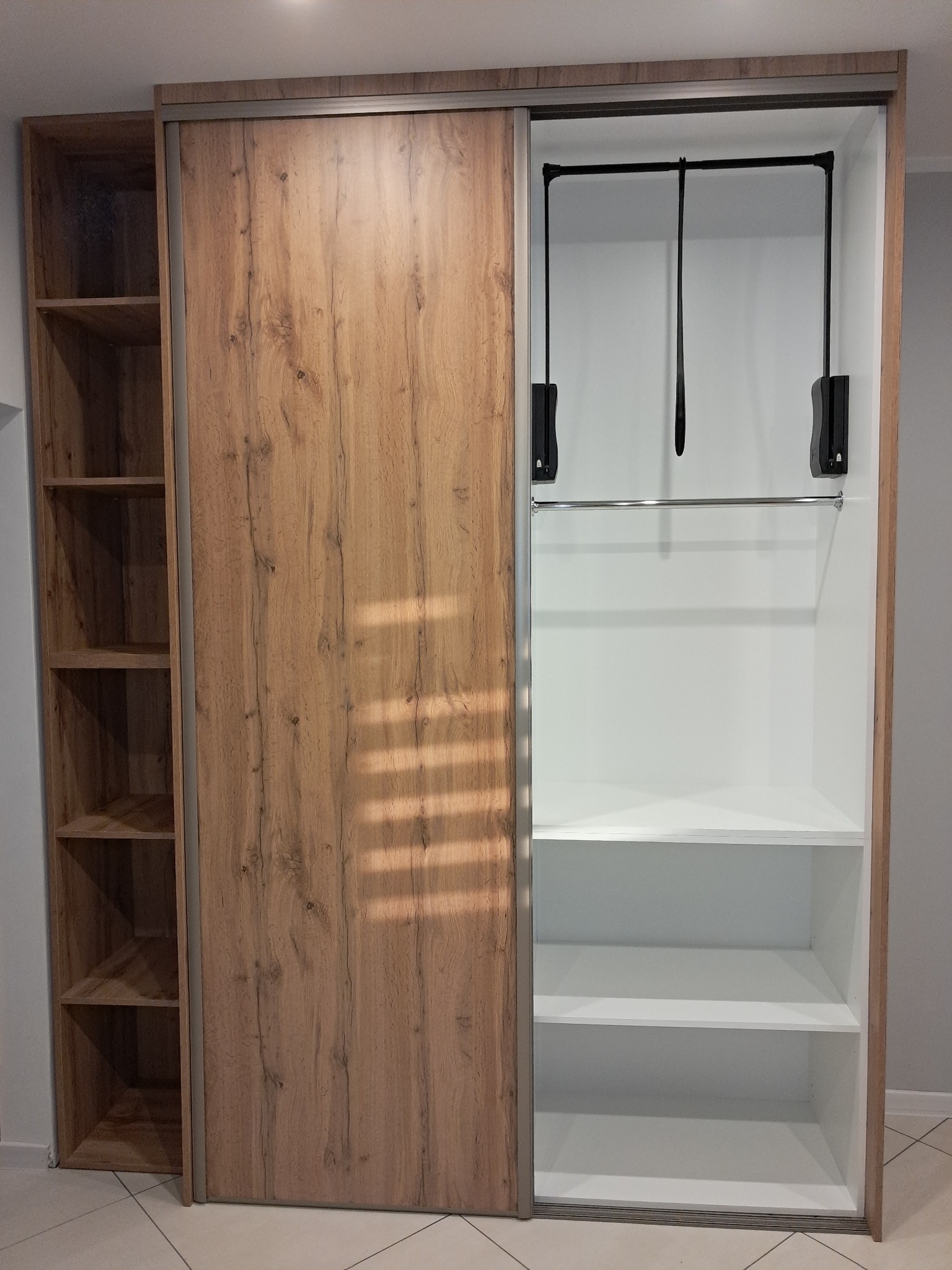 Szafa wnękowa z przesuwnymi drzwiami o teksturze drewna, z widocznym wnętrzem z półkami i drążkiem na ubrania, wykończona w neutralnej kolorystyce.