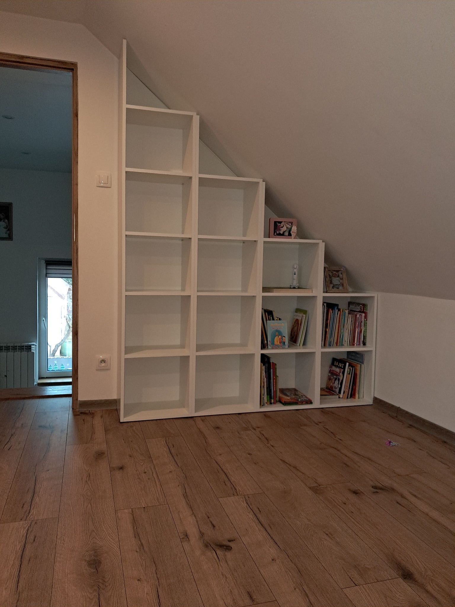 Biała, modułowa szafa wnękowa na poddaszu, z książkami i dekoracjami. Drewniana podłoga, widoczne gniazdko elektryczne i fragment drzwi. Ukośny sufit.