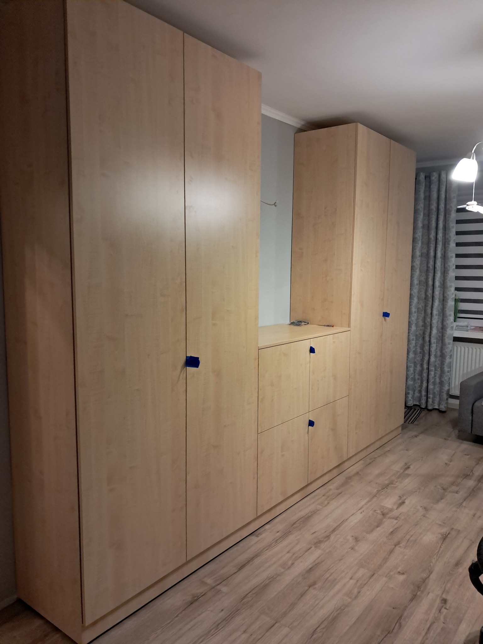 Jasna, drewniana szafa wnękowa z niebieskimi zabezpieczeniami na frontach szuflad i drzwi, ustawiona w pokoju na drewnianej podłodze.
