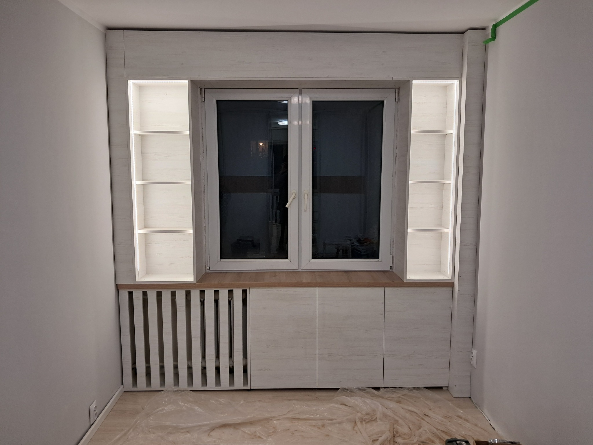 Biała zabudowa okienna z szafkami i półkami z podświetleniem LED, maskująca grzejnik, z delikatnym wzorem drewna. Element dekoracyjny wnętrza.