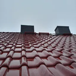 Błyszczący, bordowy dach z dachówki ceramicznej, dwa kominy wentylacyjne z czarnej dachówki, widok z dołu na pochmurne niebo po deszczu.