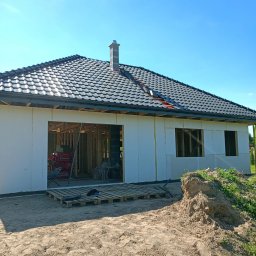 REMI-DEK PIOTR FRĄCKIEWICZ - Dom jednorodzinny w trakcie budowy, widoczny dach z ciemnej dachówki, ściany ocieplone styropianem, otwory okienne i drzwiowe, palety na ziemi.