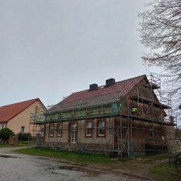 REMI-DEK PIOTR FRĄCKIEWICZ - Dom w rusztowaniach z częściowo wymienionym czerwonym dachem z dachówki. Widoczny pracownik w odblaskowej kamizelce. Pochmurne niebo.