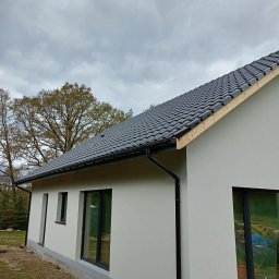 REMI-DEK PIOTR FRĄCKIEWICZ - Nowoczesny dom jednorodzinny z grafitową dachówką ceramiczną i czarnymi rynnami, widok z boku na tle pochmurnego nieba i drzew. Częściowo widoczna więźba.