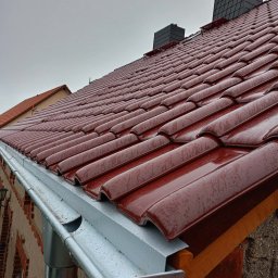 REMI-DEK PIOTR FRĄCKIEWICZ - Czerwona dachówka ceramiczna, mokra od deszczu, z zamontowanymi srebrnymi rynnami. Widok na kominy i fragment sąsiedniego dachu w pochmurny dzień.