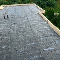 REMI-DEK PIOTR FRĄCKIEWICZ - Przygotowany płaski dach z papą termozgrzewalną i drewnianą obróbką krawędziową, widok z góry na tle zieleni i zabudowań.