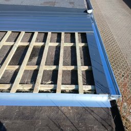 REMI-DEK PIOTR FRĄCKIEWICZ - Fragment dachu płaskiego w trakcie naprawy, z widoczną drewnianą konstrukcją pod poszyciem i nową, błyszczącą obróbką blacharską przy krawędzi dachu.