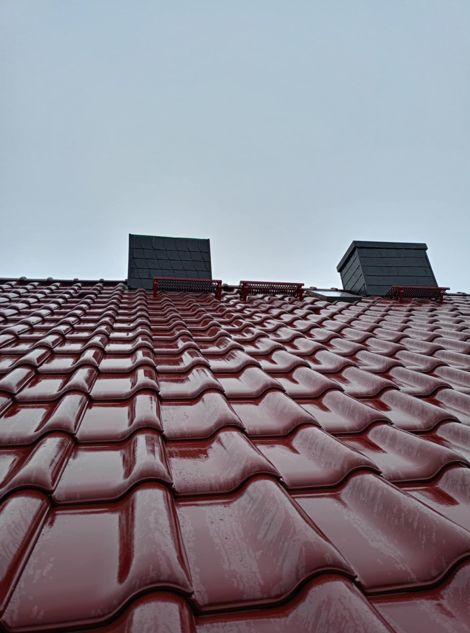 Błyszczący, bordowy dach z dachówki ceramicznej, dwa kominy wentylacyjne z czarnej dachówki, widok z dołu na pochmurne niebo po deszczu.