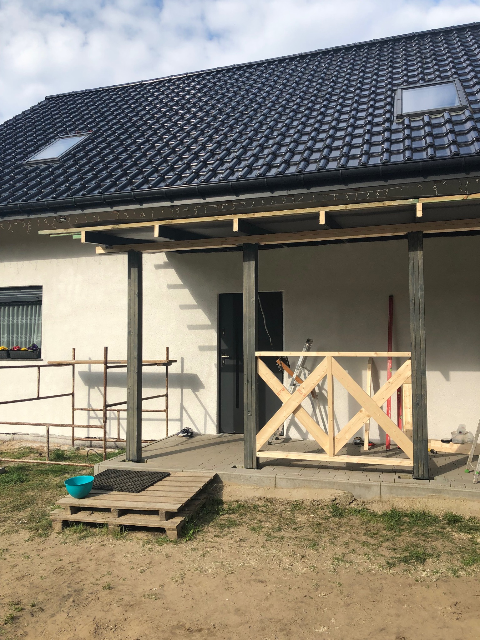 Dom z czarną dachówką i lukarnami w trakcie budowy ganku. Widoczne drewniane elementy konstrukcyjne, rusztowanie i narzędzia. Prace w toku, słoneczny dzień.