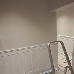 Verto - Świeżo pomalowane wnętrze z białą boazerią i otwartą skrzynką elektryczną. Aluminiowa drabina stoi w rogu, sugerując trwające prace wykończeniowe.