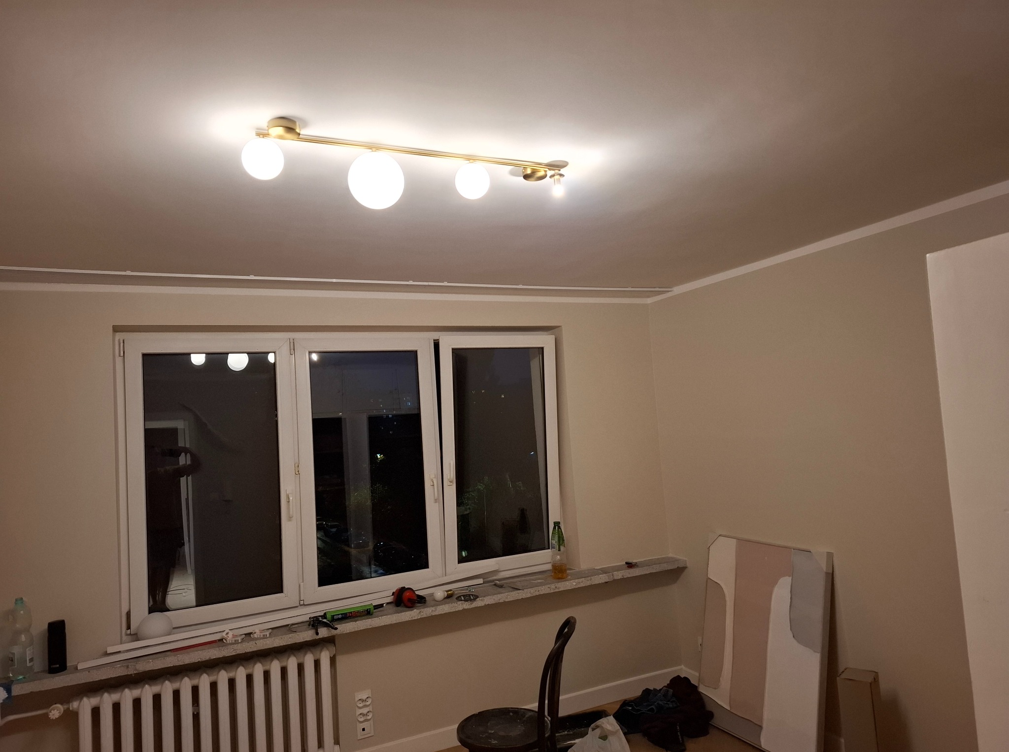 Jasne wnętrze po malowaniu: beżowe ściany, białe listwy przypodłogowe i okno z widokiem na nocne miasto. Na parapecie narzędzia malarskie.