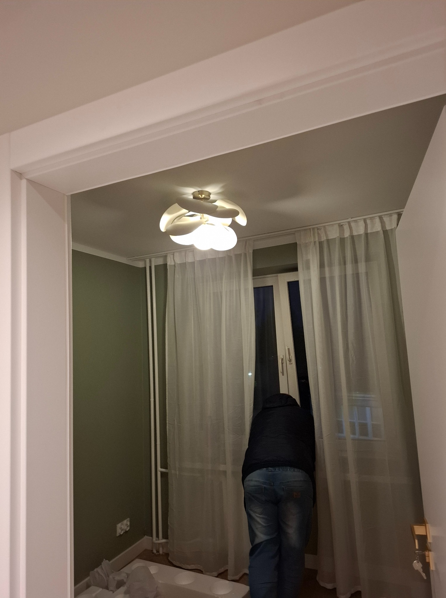Pomalowany pokój z oliwkową ścianą, białym sufitem i karniszem. Osoba w jeansach i kurtce stoi przy oknie z białymi zasłonami. Widoczny fragment drzwi i ozdobna lampa.