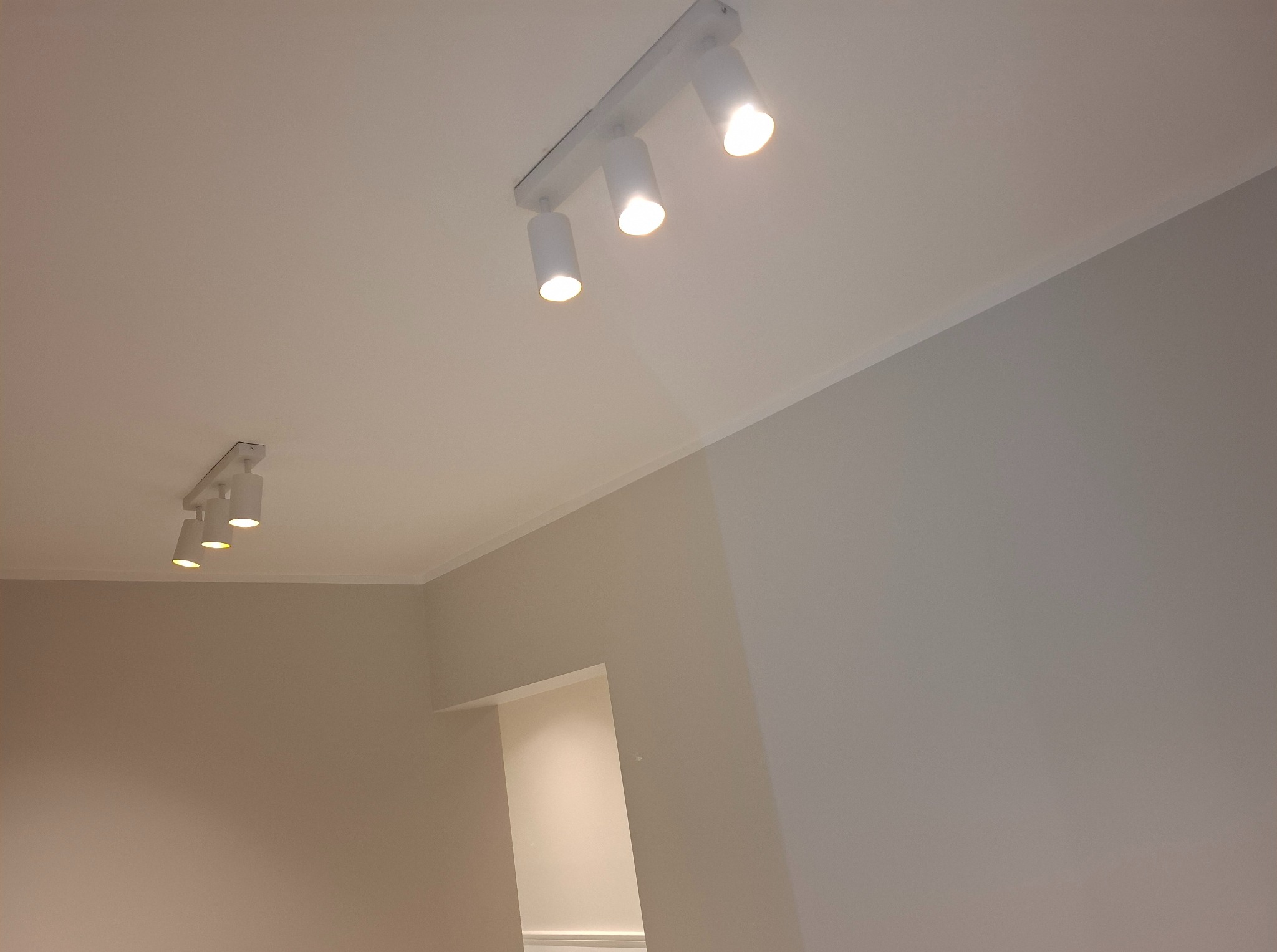 Jasne, świeżo pomalowane wnętrze z dwoma lampami sufitowymi. Ściany w odcieniach beżu i szarości, subtelny detal wykończeniowy przy suficie. Minimalistyczny design.