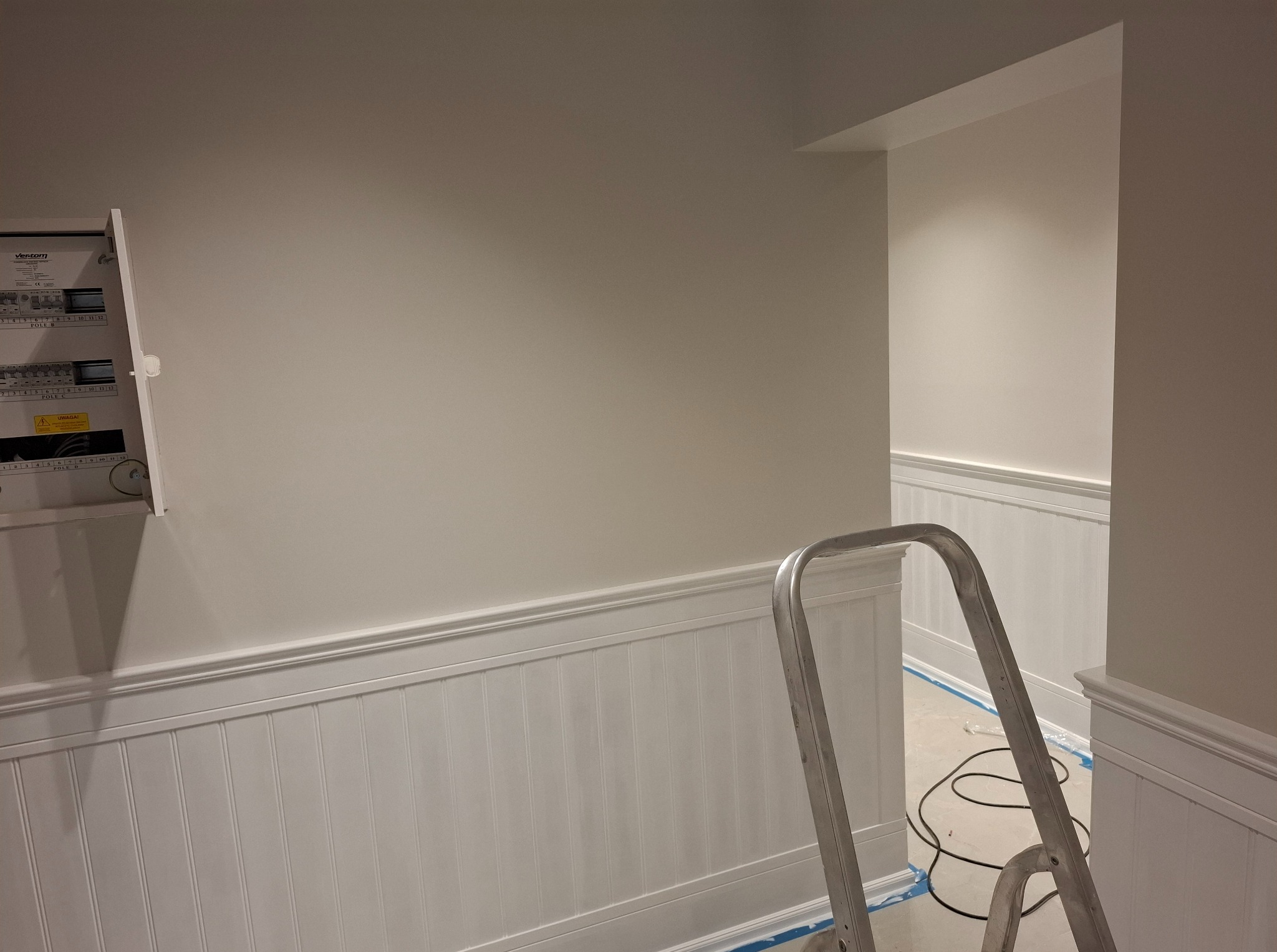 Świeżo pomalowane wnętrze z białą boazerią i otwartą skrzynką elektryczną. Aluminiowa drabina stoi w rogu, sugerując trwające prace wykończeniowe.