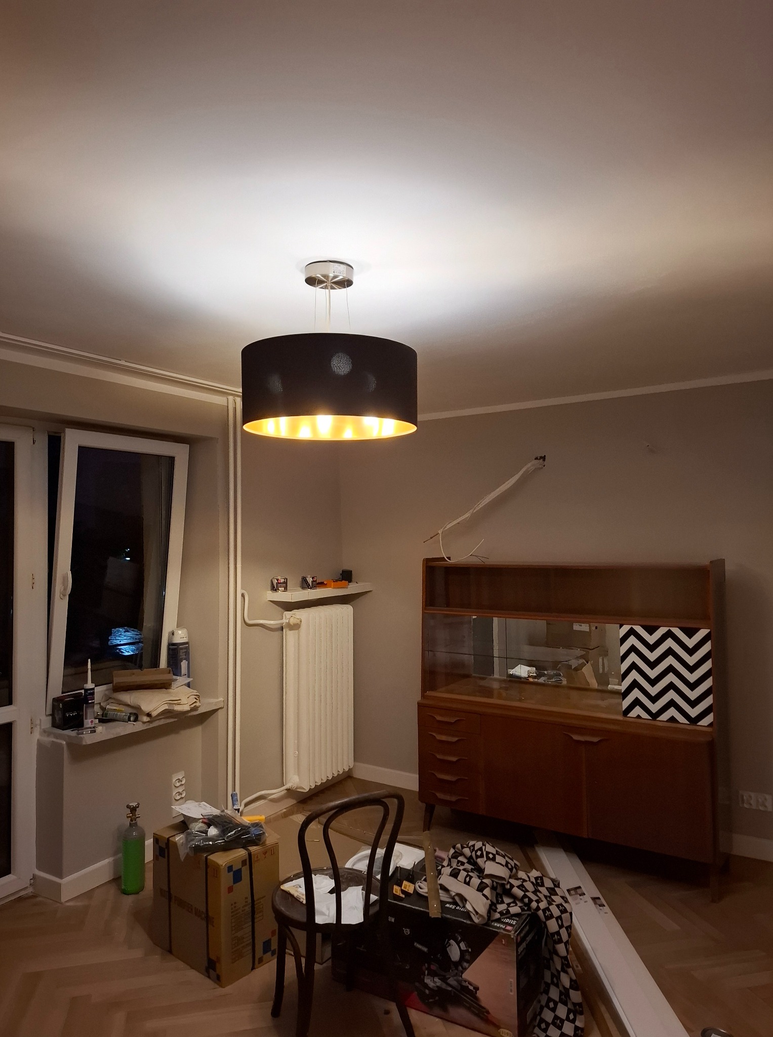 Pokój w trakcie odświeżania: ściany w neutralnym odcieniu, wisząca lampa, meble w stylu vintage i narzędzia malarskie w kartonach na podłodze.