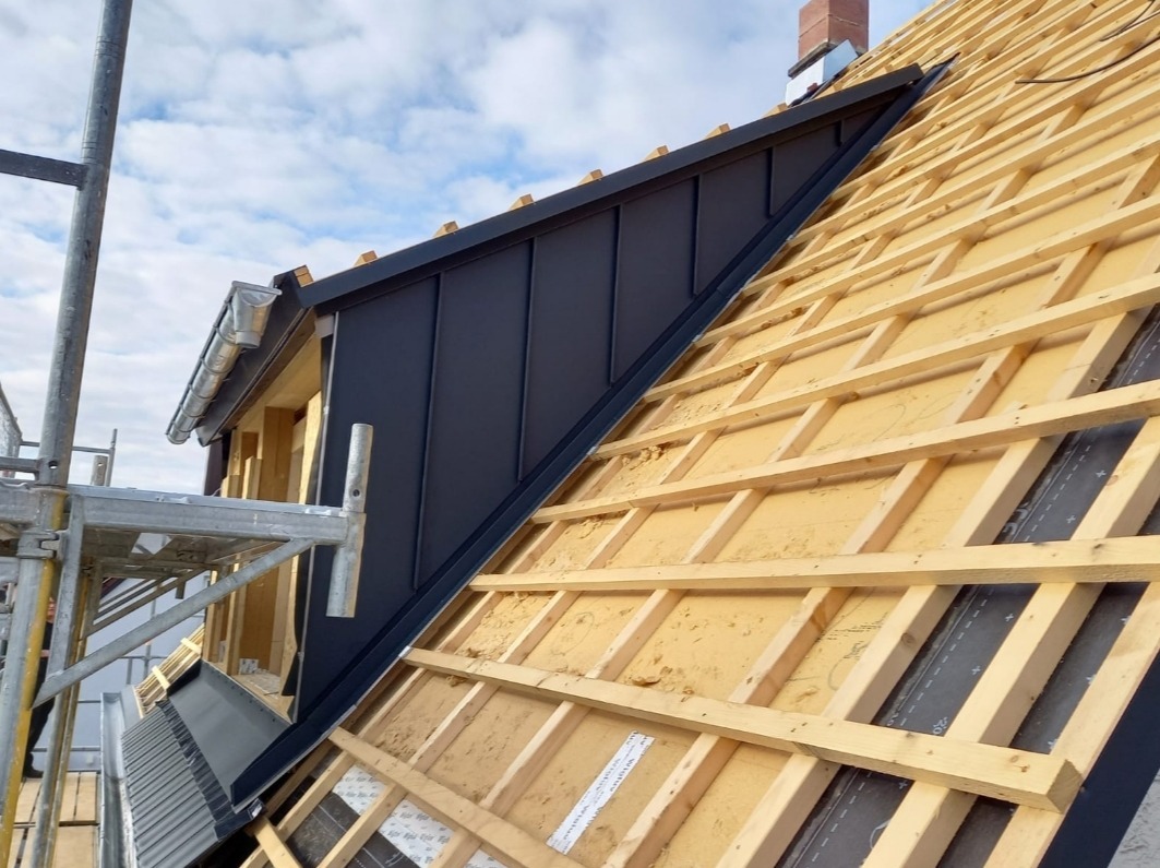 Konstrukcja dachu z deskowaniem i fragmentem czarnej blachy trapezowej; widoczne rusztowanie i rynna. Etap prac dekarskich w słoneczny dzień.