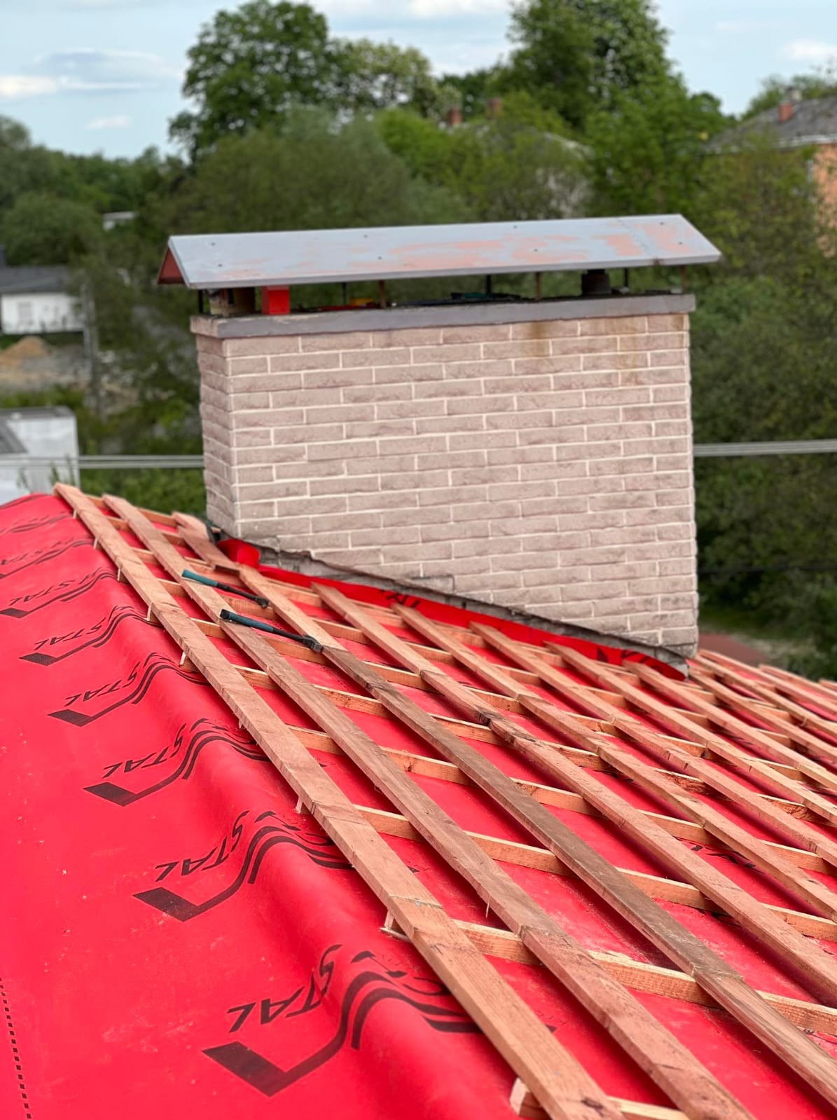 Czerwona membrana dachowa z łatami drewnianymi, komin z cegły na tle zieleni. Etap krycia dachu z bliska, widoczne narzędzia.