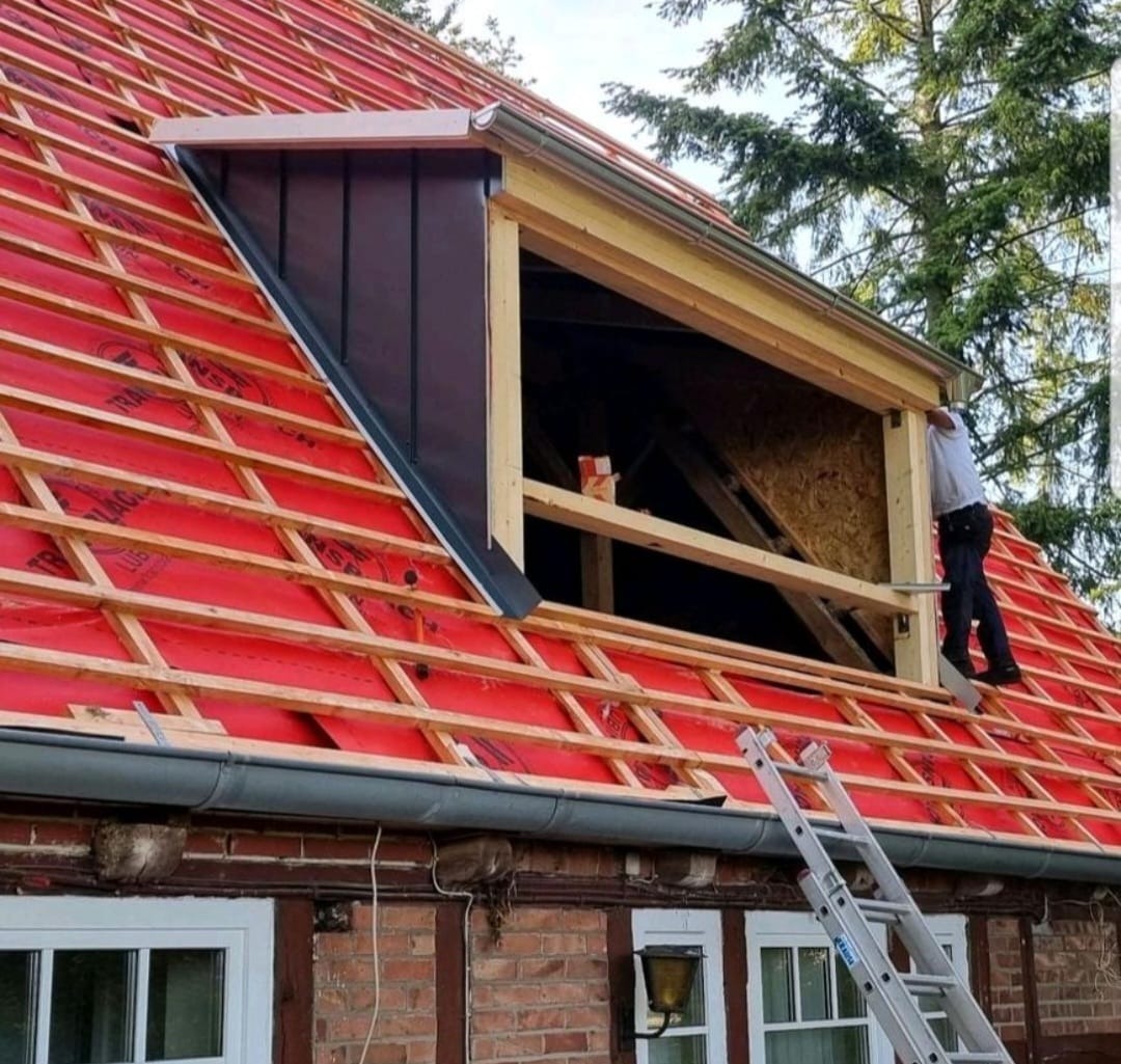 Dach z czerwoną membraną, drewnianą konstrukcją i oknem dachowym w trakcie montażu; widoczna drabina i fragment budynku z cegły.