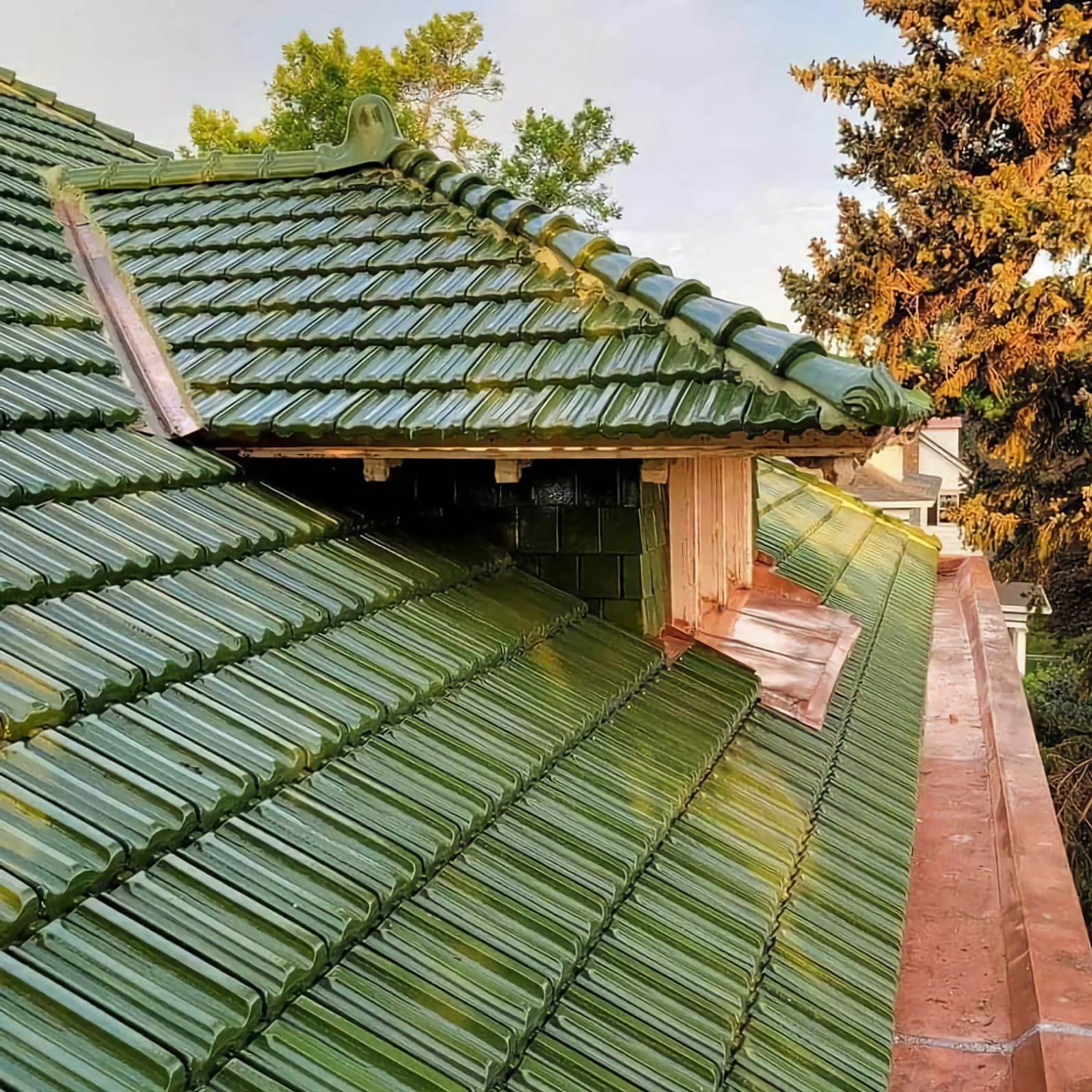 Błyszczący, zielony dach z ceramicznej dachówki karpiówki, z dekoracyjnym wykończeniem kalenicy i miedzianymi obróbkami blacharskimi, w tle drzewa i fragment budynku.