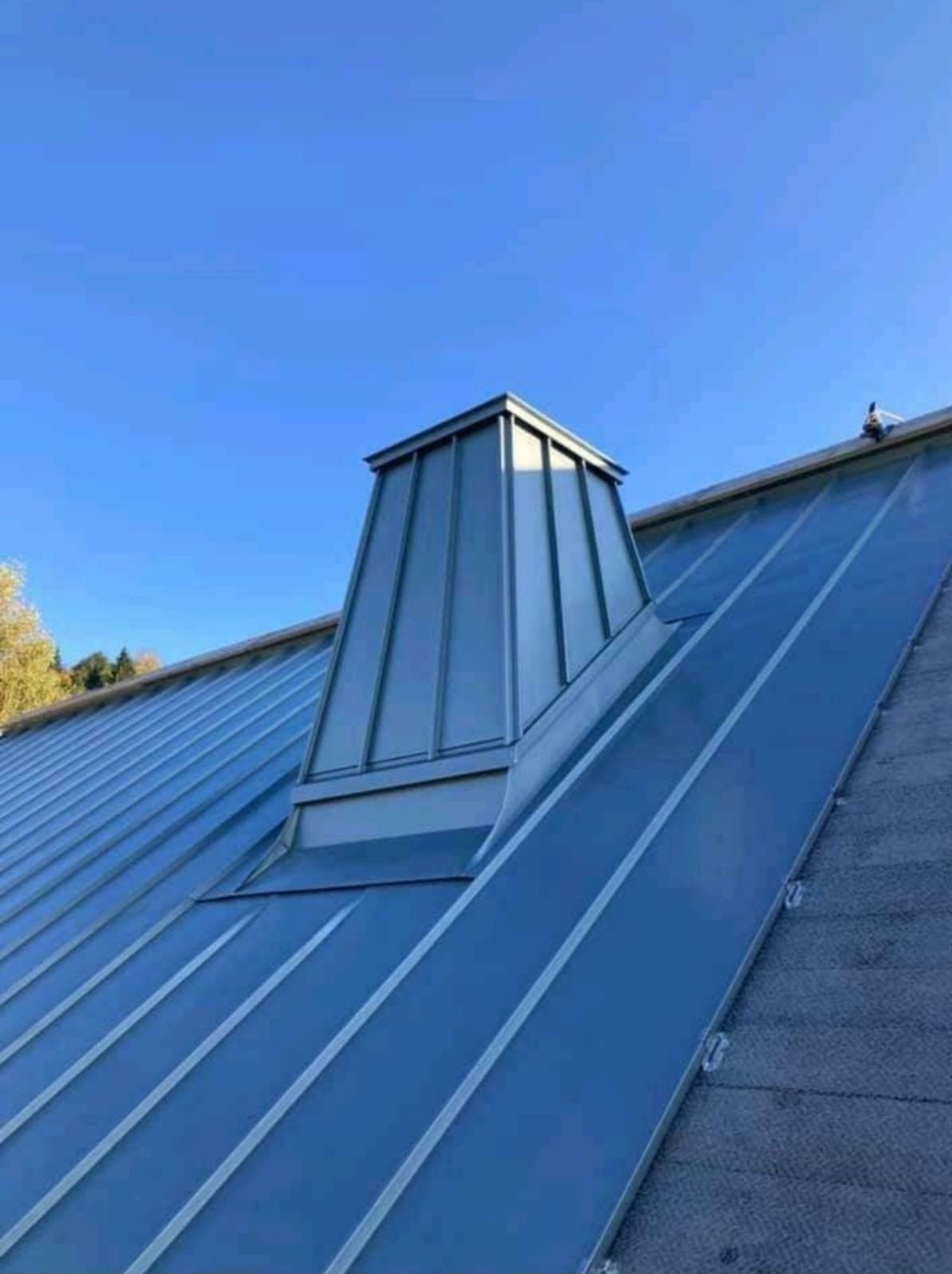 Metalowy dach z lukarną w kształcie ściętego ostrosłupa, wykonany z blachy w odcieniach szarości i błękitu, na tle błękitnego nieba. Widok z dołu, z lekkim kątem.