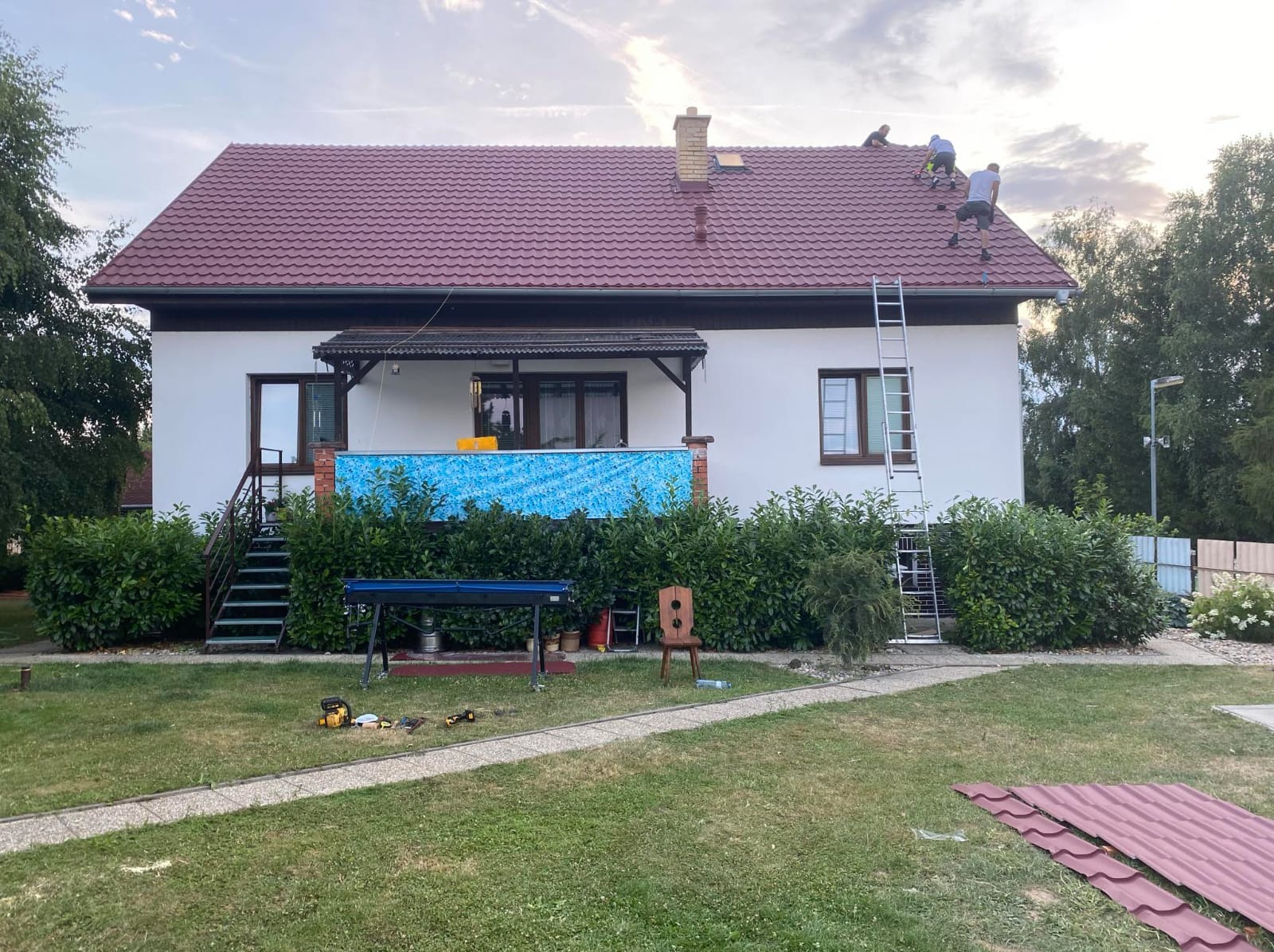 Ekipa na dachu domu z czerwoną dachówką, drabina oparta o ścianę. Na trawniku narzędzia i blacha. Prace dekarskie w toku, dom w otoczeniu zieleni.