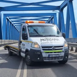 Biała laweta na moście z niebieską konstrukcją kratownicową w tle, z napisem 'MOTOJAMA Pomoc Drogowa' i pomarańczowymi światłami ostrzegawczymi.