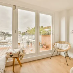 Jasne wnętrze z dużymi, przesuwnymi drzwiami balkonowymi z widokiem na taras z meblami i roślinami. Naturalne światło podkreśla drewniane podłogi.