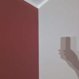 Tomasz kania - Pomieszczenie po malowaniu: ściana w odcieniu ceglanej czerwieni sąsiaduje z jasnoszarą ścianą, widoczny cień osoby robiącej zdjęcie telefonem. Wykończenie sufitu białą listwą.