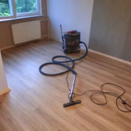 Pok&oacute;j po ułożeniu paneli podłogowych. Widoczny odkurzacz przemysłowy. Jasne panele ułożone r&oacute;wnolegle do okna. Część ściany pomalowana na szaro.