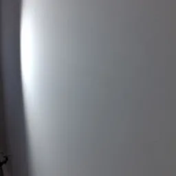 Gładka, świeżo pomalowana ściana w odcieniach bieli, z subtelnym cieniowaniem światła z lewej strony. Widoczny fragment podłogi w lewym dolnym rogu.