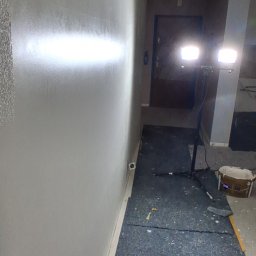 Tomasz kania - Świeżo pomalowana, gładka ściana w jasnym odcieniu szarości, oświetlona lampami budowlanymi. Na podłodze widoczne ślady farby i zabezpieczenie podłoża.