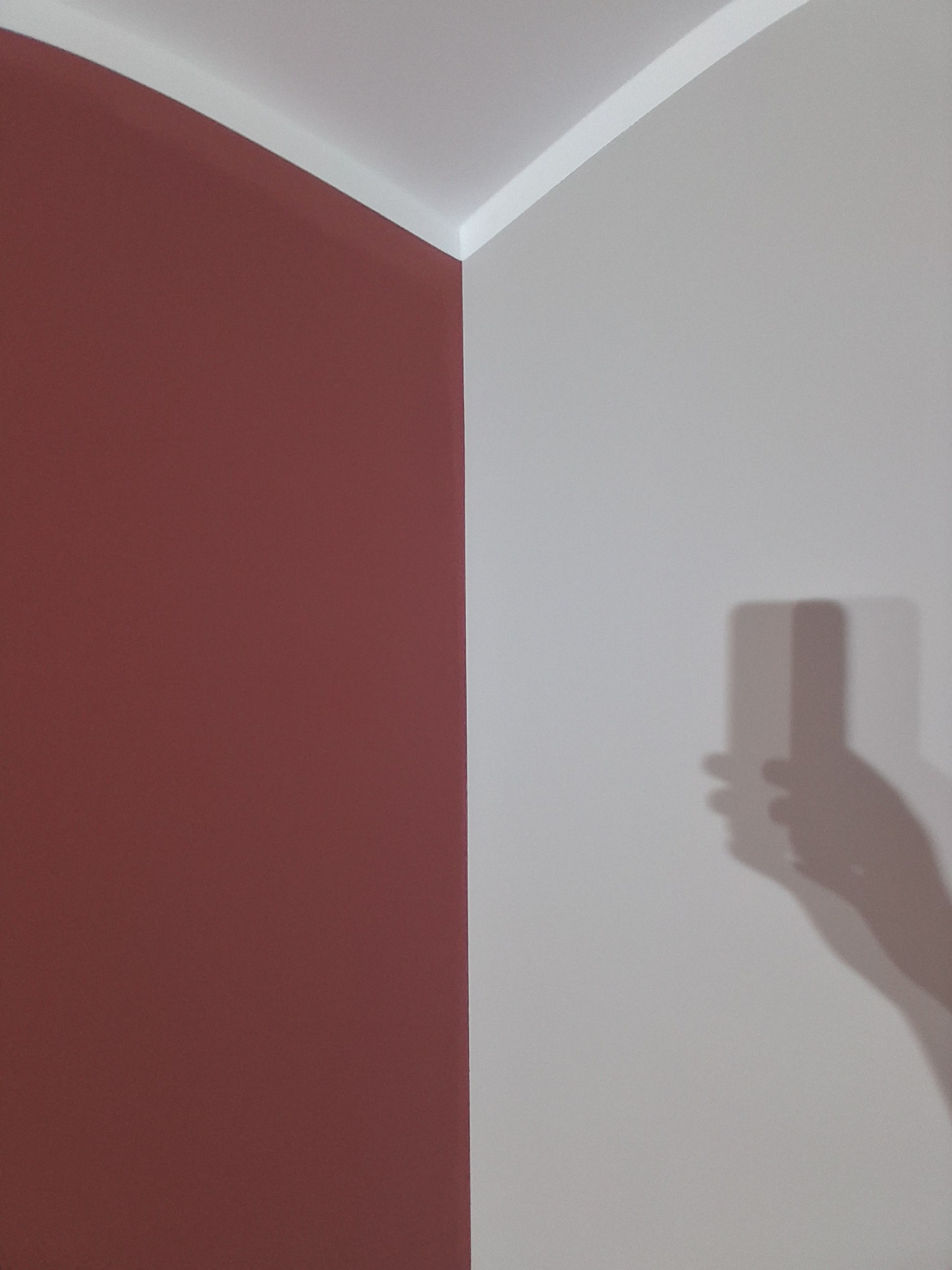 Pomieszczenie po malowaniu: ściana w odcieniu ceglanej czerwieni sąsiaduje z jasnoszarą ścianą, widoczny cień osoby robiącej zdjęcie telefonem. Wykończenie sufitu białą listwą.