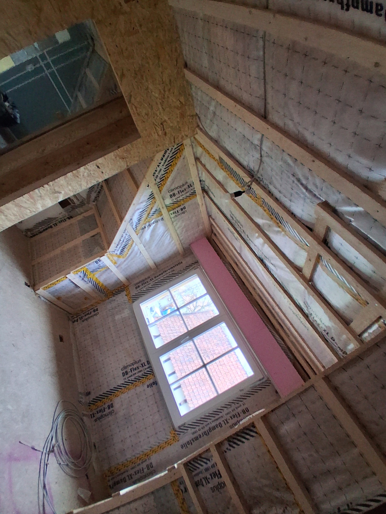 Poddasze w trakcie prac: drewniana konstrukcja szkieletowa, folia paroizolacyjna z nadrukiem, okno dachowe. Widoczne przewody elektryczne.