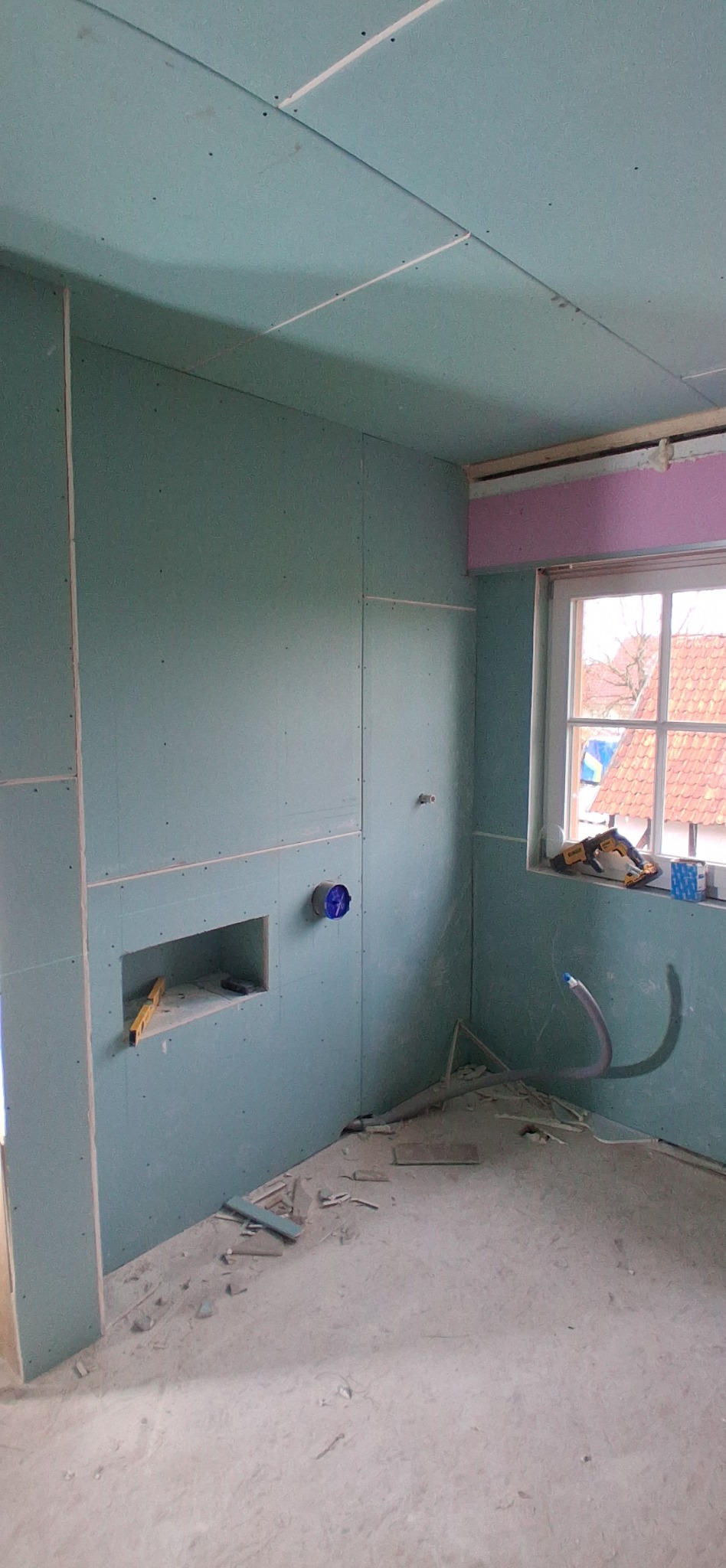 Montaż płyty karton-gips w pomieszczeniu. Widoczna wnęka w ścianie z poziomicą, instalacja wentylacyjna i narzędzia na parapecie. Ściany i sufit pokryte płytami zielonego koloru.