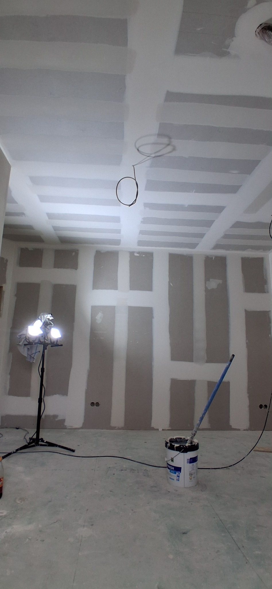 Wnętrze w trakcie prac: ściany z płyt gipsowo-kartonowych, częściowo wyszpachlowane, oświetlenie budowlane i wiadro z farbą na podłodze.