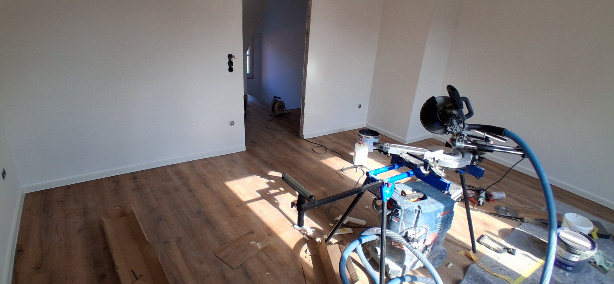 Pokój po malowaniu ścian i ułożeniu paneli podłogowych. Widoczna piła do paneli, puszki z farbą i listwy przypodłogowe. W tle widoczne kolejne pomieszczenie.