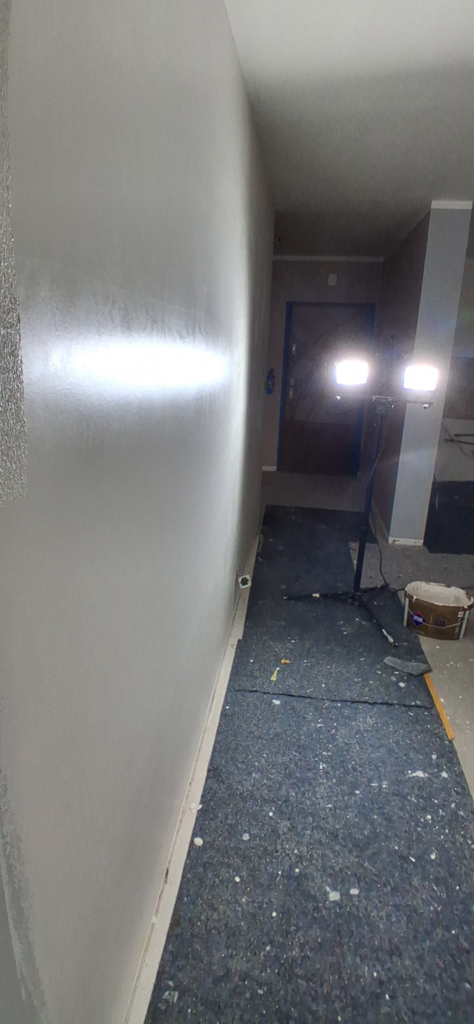 Świeżo pomalowana, gładka ściana w jasnym odcieniu szarości, oświetlona lampami budowlanymi. Na podłodze widoczne ślady farby i zabezpieczenie podłoża.