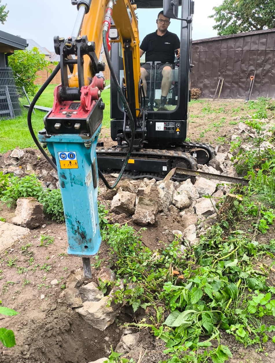 Żółta minikoparka z operatorem kruszy gruz w wykopie na działce. Widoczny gruz, zieleń i ogrodzenie w tle.