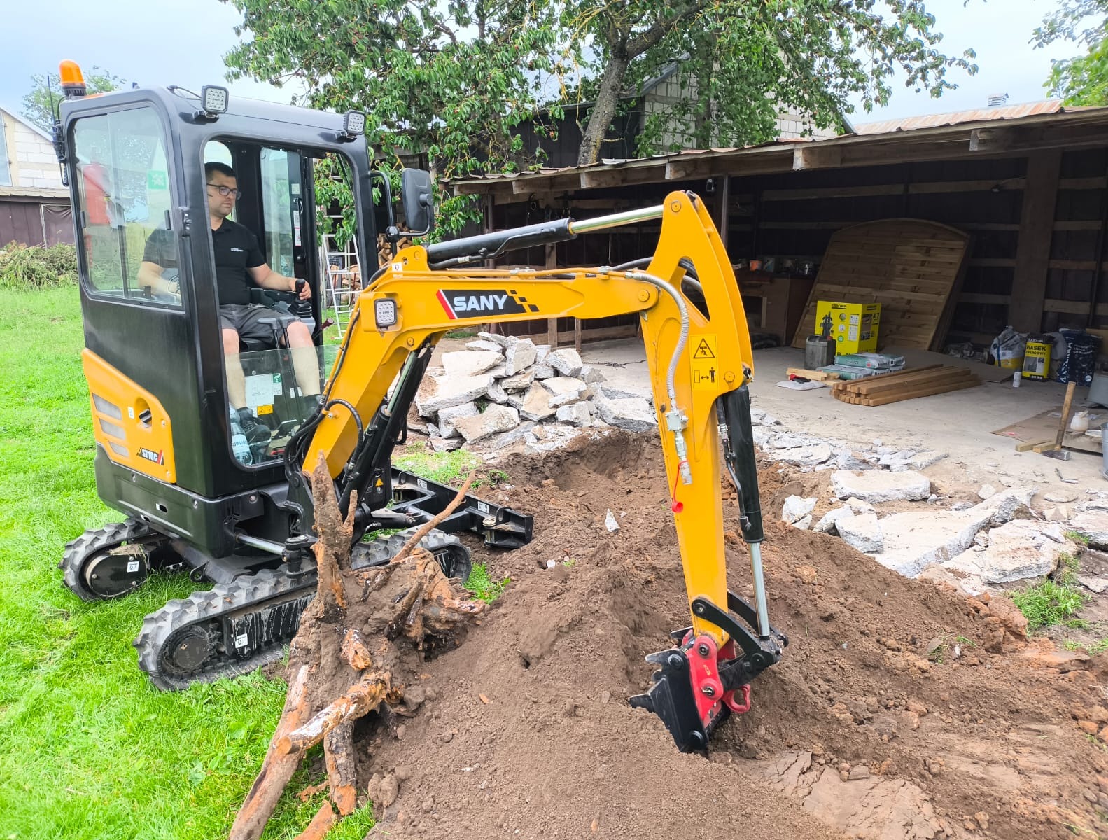 Żółto-czarna mini koparka SANY w trakcie pracy: operator w kabinie, ramię koparki zagłębia się w ziemię, obok leżą korzenie drzewa. W tle budynek gospodarczy.